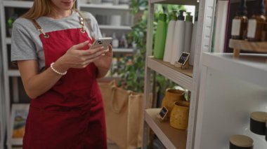 Kadın, çeşitli dekorasyon ürünleriyle çevrili telefonuna bakarken bir dekor mağazasında alışveriş yapıyor.