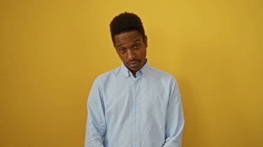 Mavi gömlekli genç adam, izole edilmiş sarı arka plan duvarına karşı duruyor, büyüleyici bir ifadeyle kendine güvenini ve tarzını aktarıyor..