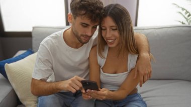 Kanepede gülümseyen bir çift, ev ortamında telefonu paylaşan bir erkek ve bir kadın, sıcak, doğal bir ışıkla bir apartman dairesinde sevgi ve mutluluk taşıyorlar.