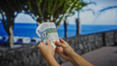 Deniz kenarında Hong Kong dolarlarını tutan eller, deniz kıyısındaki bir kasabada rahat bir yaz tatili atmosferini resmediyor, finansal yatırım ya da iş seyahati öneriyor..