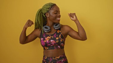 Sarı arka planda örgülü saçlarıyla güçlü bir jest yapan kadın, spor kıyafetler giyiyor, boynuna kulaklık takarak kendine güven ve neşe saçıyor..