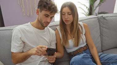 Çift rahat bir oturma odasında kanepede oturuyor, kadın ve erkek birlikte akıllı telefona bakıyor, sıcak ve samimi bir ev ortamı yaratıyor..