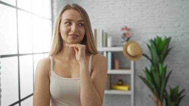 Evin içinde gülümseyen bir kadın, modern dekorasyon kitaplıkları ve bitki ışıkları ile parlak bir oturma odasında düşünceli bir şekilde, sakin bir ortamda, gündelik güzelliğini arttırıyor.