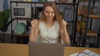 Ofis ortamında başarıyı kutlayan kadın, gülümsemesi ve yumruklarıyla, sarı saçlarıyla ve günlük kıyafetleriyle, ev dekorasyonu ve modern çalışma ortamıyla çevrili..