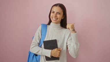 Genç kadın gülümseyen, başparmağıyla işaret eden, pembe duvarın üzerinde izole edilmiş bir kitap tutan.