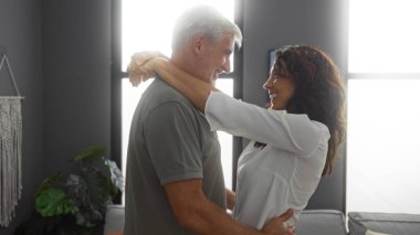 Modern oturma odasında kucaklaşan orta yaşlı bir çift, sıcak, neşeli ifadeleri ile kapalı mekanlarda sevgi ve mutluluğu yakalıyorlar..