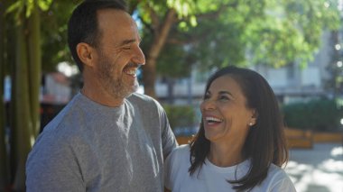 Orta yaşlı İspanyol çift, şehir parkında güneşli bir ortamda, yeşil ağaçlar ve kentsel ortamlarla çevrili mutluluk ve sıcaklığı yansıtan sevgi dolu bir anın tadını çıkarıyorlar..