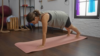 Genç bir kadın spor aletleri ve arka planda iplerle kapalı bir spor salonunda pembe bir paspas üzerinde tahta egzersizi yapıyor.