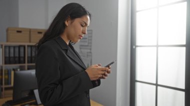 Modern ofiste siyah takım elbiseli kadın akıllı telefon kullanıyor rafları ve arka planda penceresi var..