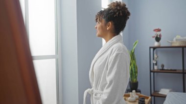 Genç bir kadın spa odasında gülümsüyor, beyaz bir bornoz giyiyor, kıvırcık saçlı ve sakin bir yüz ifadesi var. Parlak bir sağlık merkezinde pencereden dışarı bakıyor.