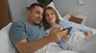 Bir çift, yatak odasında, yatak odasında, samimi bir şekilde gülümseyen bir erkekle ve bir kadınla telefonda rahatlıyor. Bu da samimiyet ve rahatlama göstergesidir..