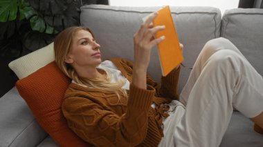 Kadın rahat bir oturma odasında, rahat bir atmosferi olan koltukta kitap okuyor, evde rahatlık ve sıcaklık sergiliyor..
