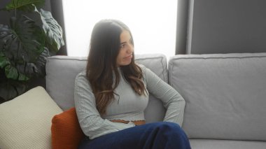 Oturma odasında rahat bir kanepede oturan bir kadın. Bitkiler ve yumuşak yastıklarla. Huzurlu ve rahat bir ev atmosferini yansıtıyor..