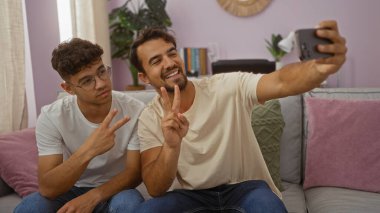 Baba ve oğul zafer işaretleri yapıyor ve rahat bir oturma odasında selfie çekiyor, bağlarını gösteriyor ve mutluluklarını paylaşıyorlar.