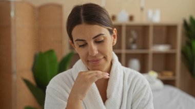 Kaplıcadaki genç İspanyol kadın, sakin bir ifadeyle bornoz giyiyor, esmer, sağlık merkezinde, kapalı bitkiler ve rahatlatıcı ambiyansı olan.