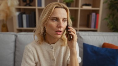 Akıllı telefondan konuşan bir kadın, rahat bir oturma odasında oturmuş, etrafı yumuşak dekorlar ve kitaplıklarla çevrili, samimi bir ev ortamının altını çizerek endişelerini dile getiriyor..