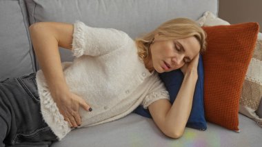 Parlak oturma odasında rahat bir kanepede uzanan bir kadın, elleri karnında rahatsızlık belirtileri gösteriyor, güzel sarışın, orta yaşlı, evde dinlenen bir yetişkin..