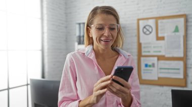 Modern ofis ortamında akıllı telefon kullanırken gülümseyen kadın, parlak ışık altında profesyonel bir ilişki ve iş ortamında iletişim gösteriyor.