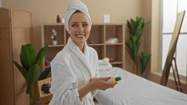 Kafasında havluyla bornoz giyen genç bir kadın bir ürünü sıcacık bir kaplıcada tutarken gülümsüyor..