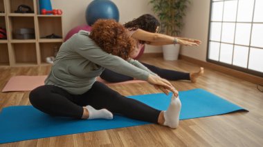 Kadınlar spor salonunda paspaslara uzanır, olgun bir dostluk ve spor rutinine olan bağlılıklarını sergilerler..