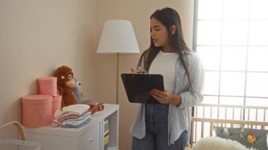 Yatak odasında pano ile duran bir kadın beşikteki bebek odasını gözetliyor, oyuncaklar ve mobilyalarla evin içini gösteriyor, şefkatli bir atmosferi resmediyor..