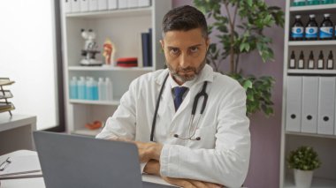 Klinikteki doktor masasında tıbbi kitaplarla ciddi görünüyor, laboratuvar önlüğü giyiyor, hastane ekipmanlarıyla çevrili, sağlık hizmetleri için çalışma ortamını ön plana çıkarıyor..