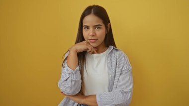 Koyu sarı arka planda poz veren, esmer saçlı, sıradan giyinmiş çekici ve kendinden emin ifadesini sergileyen bir kadın..