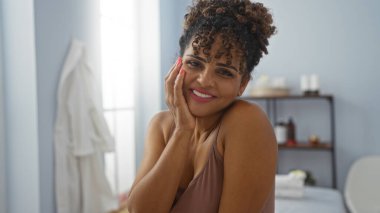 Güzeller güzeli, kıvırcık saçlı, spa odasında kameraya gülümseyen bir İspanyol kadın. Evin içinde, sağlıkla ilgili esaslarla çevrili.