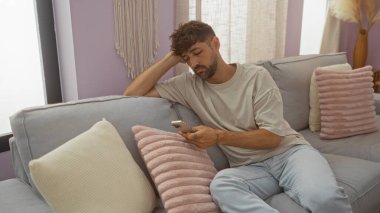 Evdeki kanepede oturan genç adam, pastel renkli yastıkları ve dekorları olan, rahat bir apartman dairesinde, düşünceli bir ifadeyle telefonuna bakıyor.