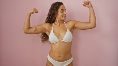 Genç esmer, İspanyol bir kadın pembe arka plan duvarının üzerinden izole edilmiş beyaz bir bikiniyle kollarını esnetiyor.