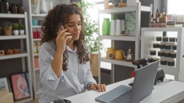 Dekoratörde telefonla konuşan bir kadın, dizüstü bilgisayarı ve etrafını dekore eden eşyalarla dolu rafları var.