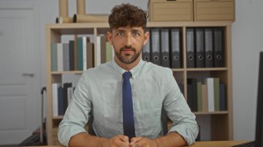 Ofiste oturan yakışıklı bir adam kravat ve gömlek giymiş, arka planda kitap ve klasörlerle dolu raflar var.