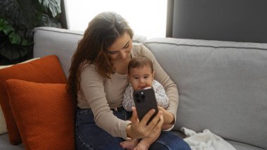 Bir kadın, küçük kızıyla rahat bir oturma odasında selfie çekiyor. Aile sevgisini sergiliyor ve koltukta akıllı telefonuyla kapalı bir evde samimi bir annelik bağı kuruyor..