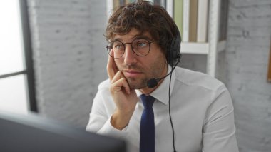 Ofis odasında kulaklık takan genç bir adam, profesyonel ortamda çalışırken düşünceli görünüyor, odaklanmış ifade ile modern iş ortamını gösteriyor..