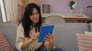 Genç bir kadın oturma odasında rahatlatıcı bir anın tadını çıkarıyor, mavi bir tablet kullanıyor, etrafı kanepe ve kitaplıkla çevrili, modern bir ev hayatı hissi uyandırıyor..