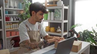 Arka planda dekoratif eşyalarla dolu raflarla dolu bir masada otururken ev dekorasyonu mağazasında çalışan sakallı bir genç.