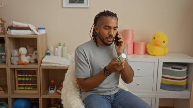Örgülü adam oturma odasında oturmuş, elinde ilaç şişesiyle telefonla konuşuyor. Etrafı oyuncak ve kitaplarla çevrili.