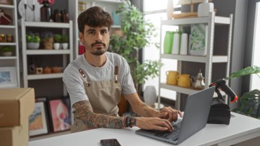 Genç, yakışıklı, sakallı, dövmeli ev dekorasyonu dükkanında dizüstü bilgisayarda çalışan, şık dekorasyon ürünleri, bitkiler ve raflarla çevrili bir adam..