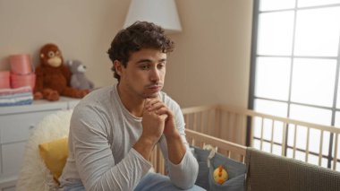 Genç İspanyol bir adam, ahşap bir bebek beşiğinin yanında, rahat bir yatak odasında, yumuşak oyuncaklar ve sıcak aydınlatmayla çevrili, bir anlık tefekkür tasvir ederek, düşünceli bir şekilde oturuyor..