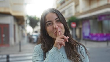 Kentsel sokak ortamında sırıtan esmer saçlı bir kadın güneşli bir şehir ortamında dikilirken suskun bir jest yapıyor gündelik ve gençlik tarzını sergiliyor..