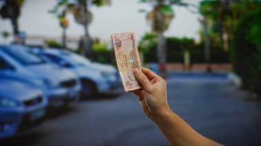 Şehir merkezinde park halindeki arabaların yanında el ele tutuşan Dirham banknotları kira veya ödeme vurgu işi ve finansal dinamik gibi işlemleri öneriyor..