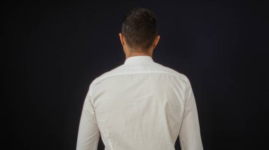 Beyaz tişörtlü, siyah arka planda, kameradan uzak duran Latin asıllı genç adam.