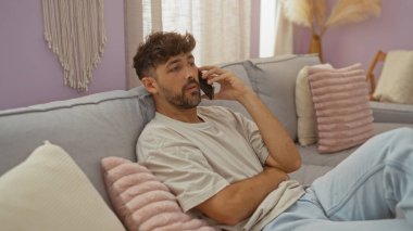 Rahat bir oturma odasında rahat rahat oturan, cep telefonuyla konuşan ve etrafı dekoratif yastıklarla ve dekoratif dekoratif malzemelerle çevrili bir koltukta rahatça oturan genç adam..