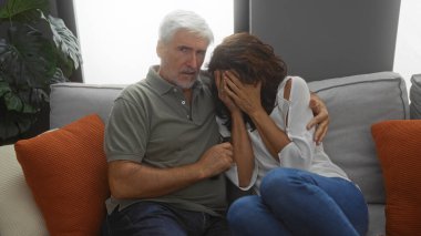 Middle aged couple watching horror movie in living room, man comforting scared woman sitting together on sofa indoors, cozy home setting suggests intimacy and shared experience. clipart