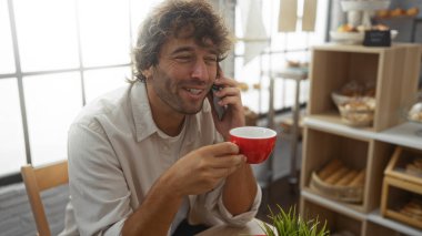 Genç adam telefonda gülümsüyor ve konuşuyor. Elinde fırında kırmızı bir kahve fincanı, arkasında da ekmek rafları var.