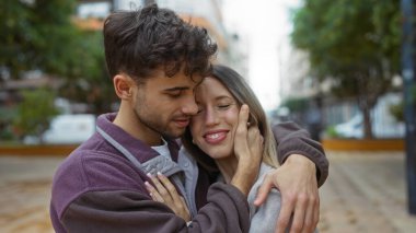 Bir şehir sokağında sevgi dolu bir şekilde kucaklaşan erkek ve kadın, açık gökyüzünün altında ilişkilerini gösteriyor ve şehir onları sıcak romantik bir anda çevrelemiş durumda..