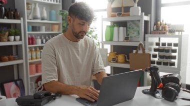 Genç adam dizüstü bilgisayarda ev dekorasyonu ve aksesuarlarıyla çevrili bir mağazada çalışıyor.