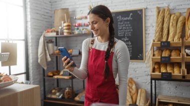 Kırmızı önlüklü genç kadın ekmek ve hamur işleriyle çevrili fırında akıllı telefon kullanıyor.