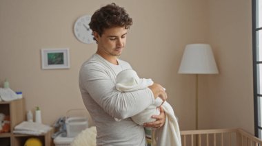 Latin kökenli genç bir adam, beşiği olan rahat bir yatak odasında bebeği tutuyor, şefkatli bir şekilde sarılıyor ve atmosferi besliyor..