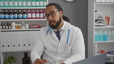 Klinikteki sakallı genç adam steteskop ve beyaz önlük giyerek tıbbi tedaviyi kendinden emin bir ifadeyle tartışıyor ve hasta bakımına ve profesyonel davranışlarına odaklanıyor..